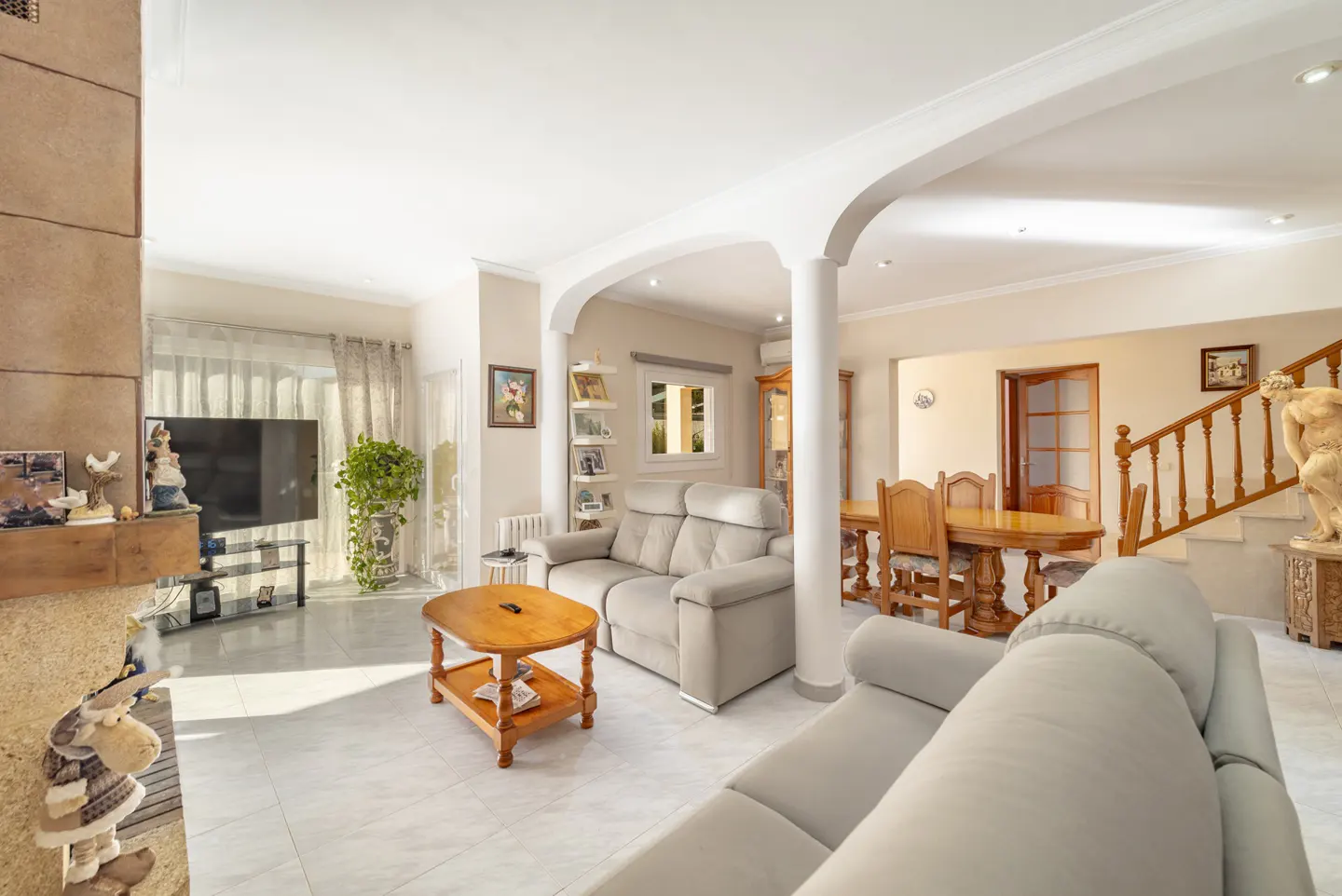 Bright, open-plan living and dining area with tile floors, beige sofas, wood table and chairs, and staircase with wood railing.