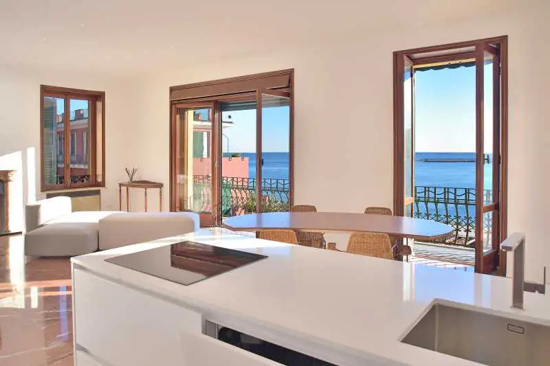 Bright, modern kitchen and dining area with ocean view. White cabinets, wood table, and wicker chairs. Balcony with blue water visible.