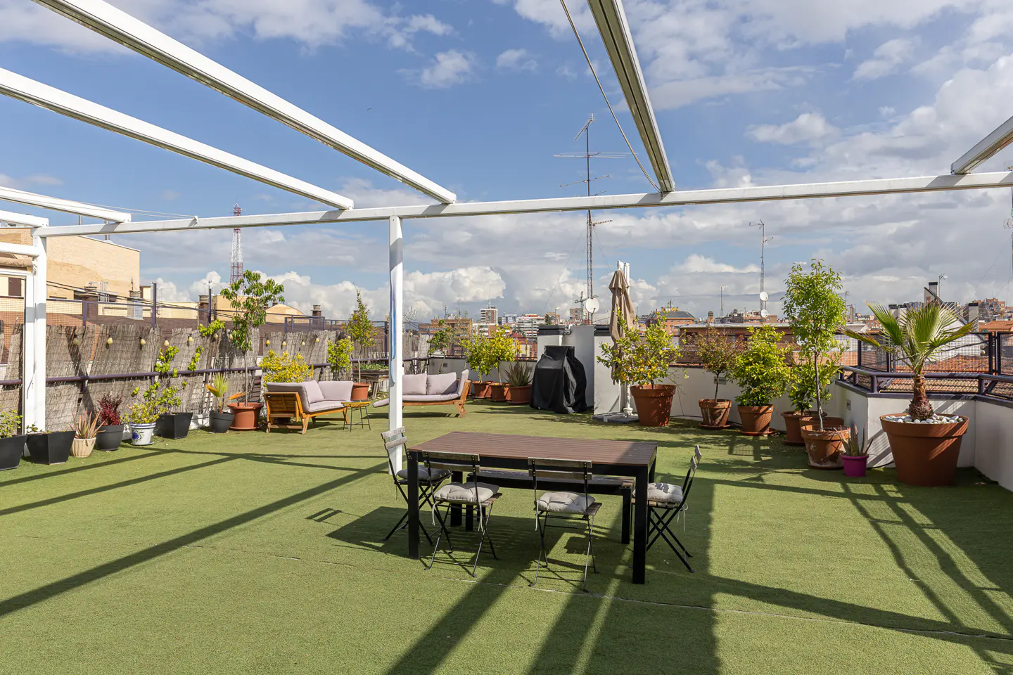 Rooftop terrace with green turf, dining table, chairs, sofas, potted plants, and a city view under a partly cloudy sky.