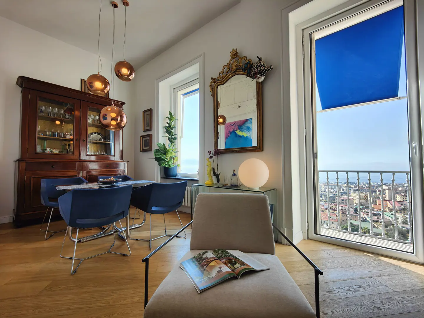 Bright living room with a dining table, wooden cabinet, and a chair with a magazine. A large window shows a city view.