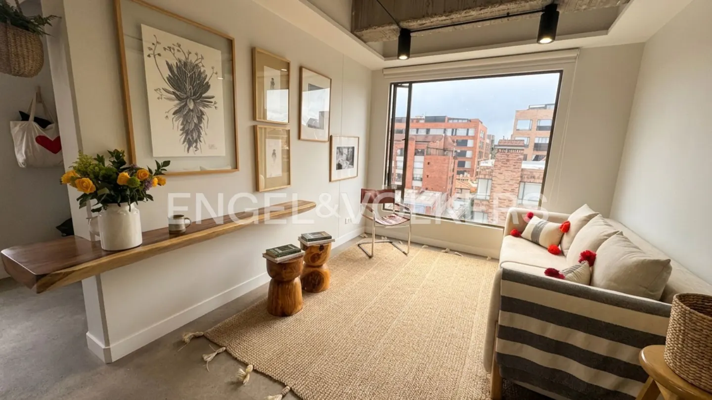 Bright living room with a beige sofa, jute rug, and a large window overlooking a city. Artwork and flowers decorate a wooden shelf.