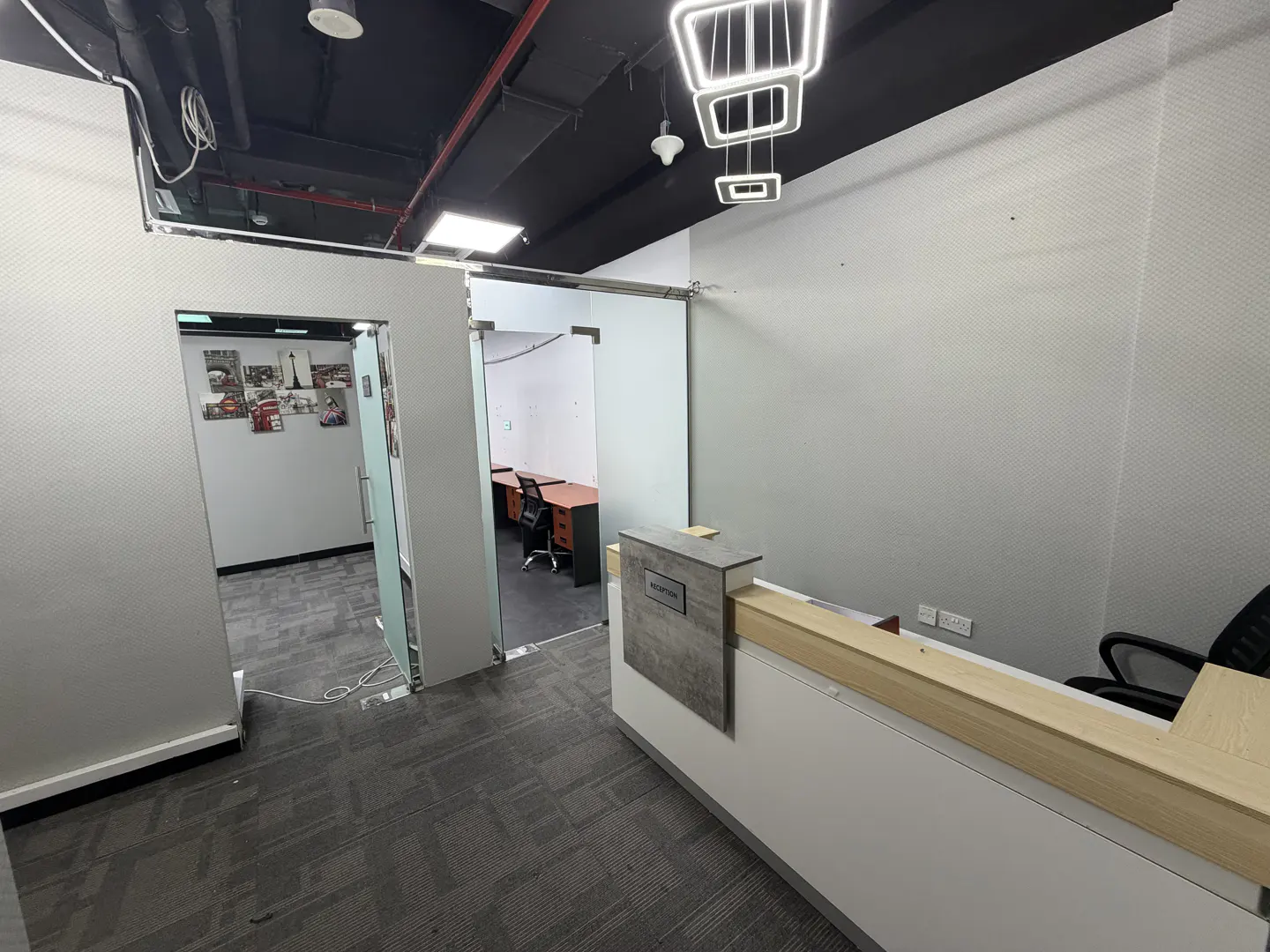Office space with a reception desk, gray carpet, and glass-walled offices. A modern light fixture hangs from the black ceiling.