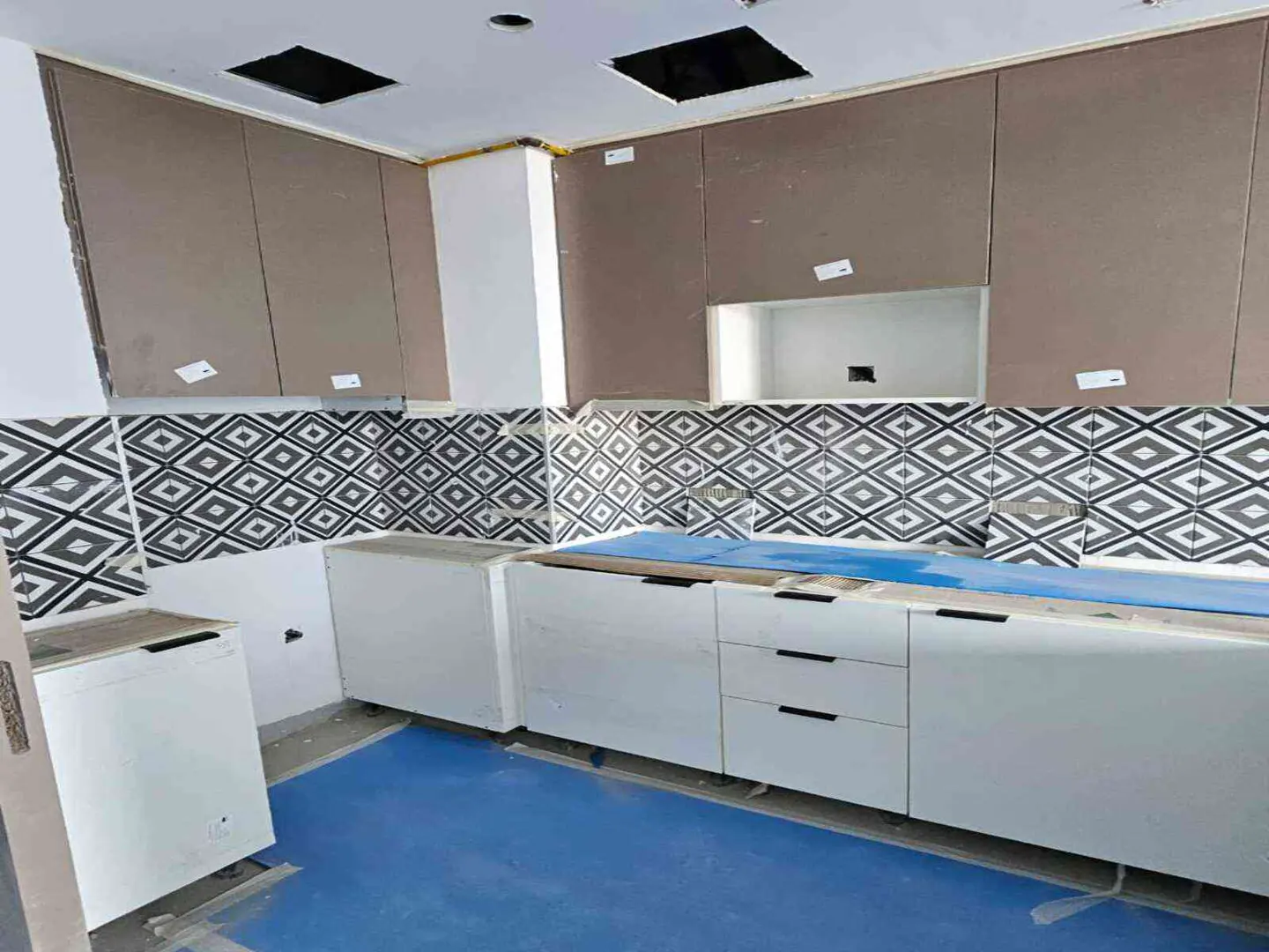 Unfinished kitchen with white cabinets, black drawer pulls, and diamond patterned backsplash. Brown cabinets above. Blue floor covering.
