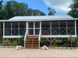 Exterior view of a white, elevated cabin with a screened porch and wooden stairs. Green trees and a blue sky are in the background.