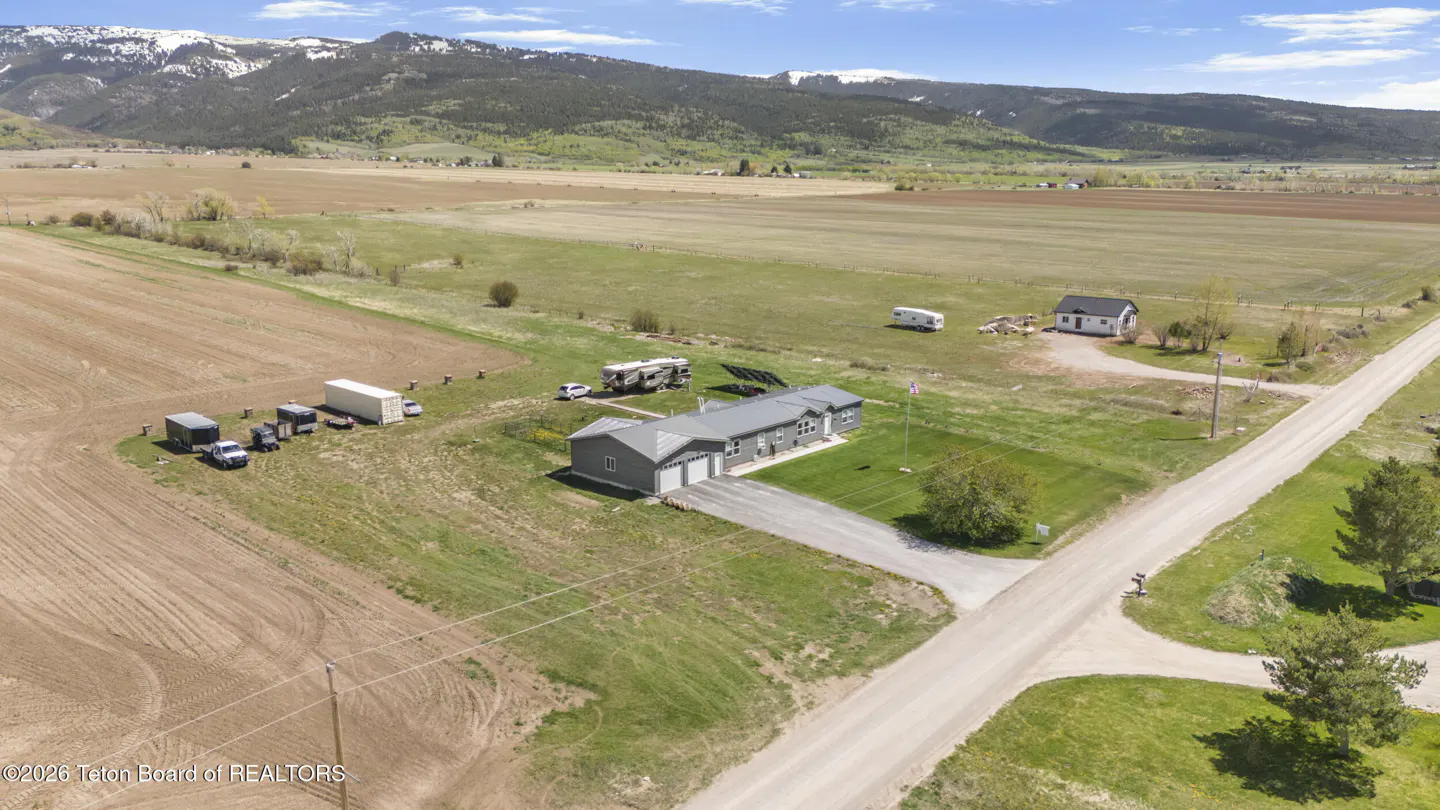 Aerial view of a gray house with a two-car garage, surrounded by green grass, fields, and mountains in the background.