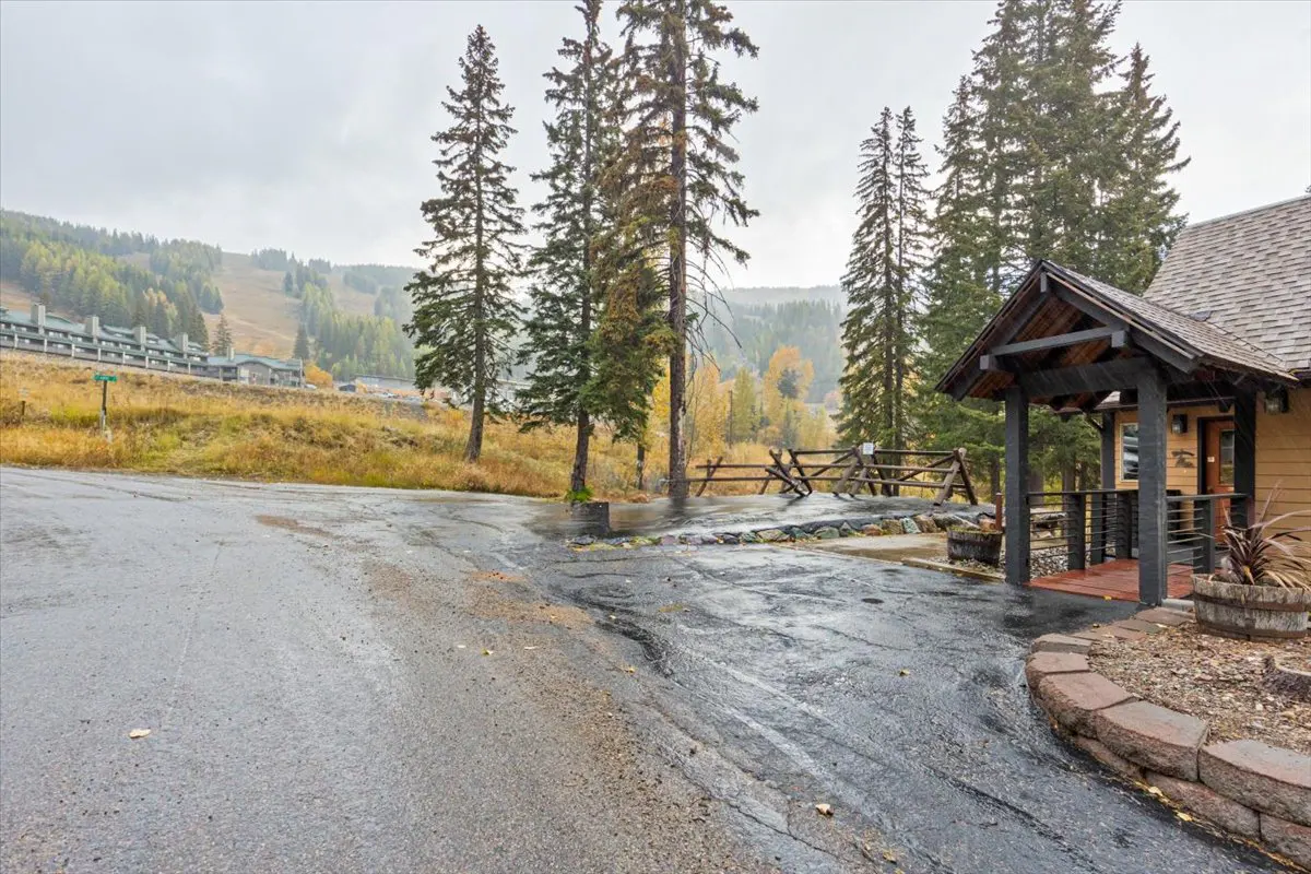 Exterior view of a mountain home with a wood porch and a gray asphalt driveway on a cloudy day. Tall pine trees and a ski slope are in the background.