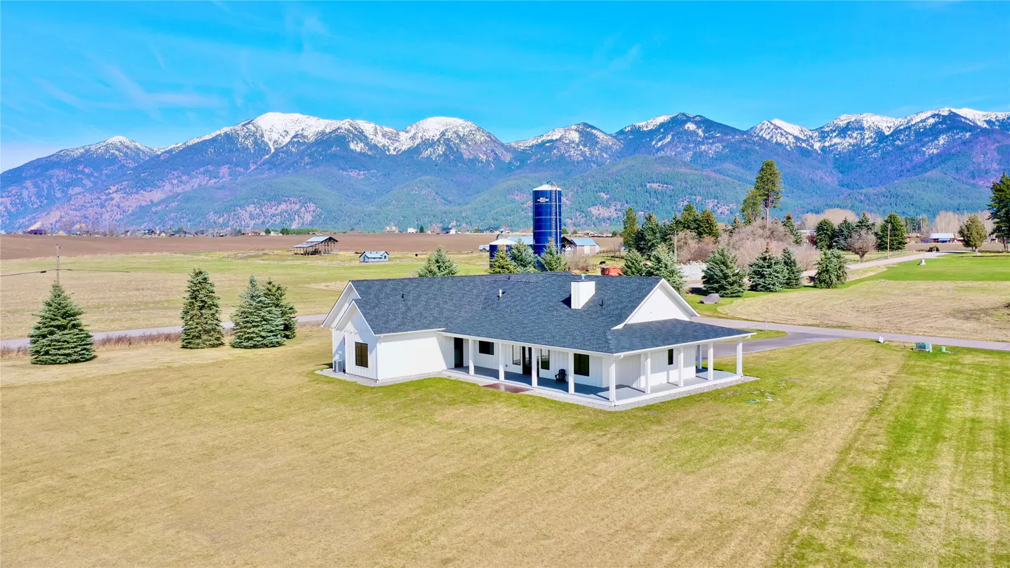 A white farmhouse with a gray roof sits in a green field with snow-capped mountains in the background.