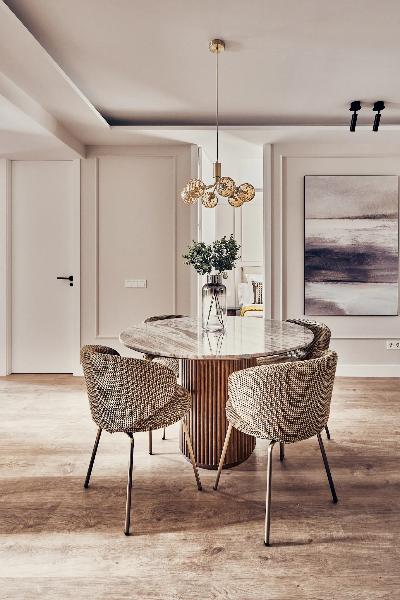 A modern dining room features a round marble table with a fluted wood base, surrounded by four woven chairs with gold legs. A gold chandelier hangs above.