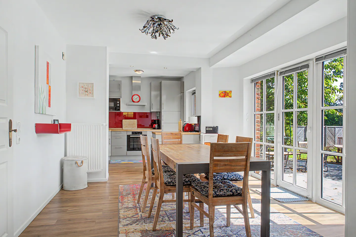 Modern dining room with a wooden table, patterned rug, and open-plan kitchen featuring a red backsplash and glass doors.