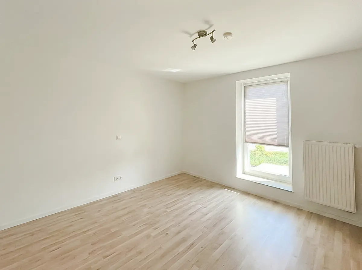Bright, empty room with light wood floors, white walls, and a window with blinds. A radiator is under the window.