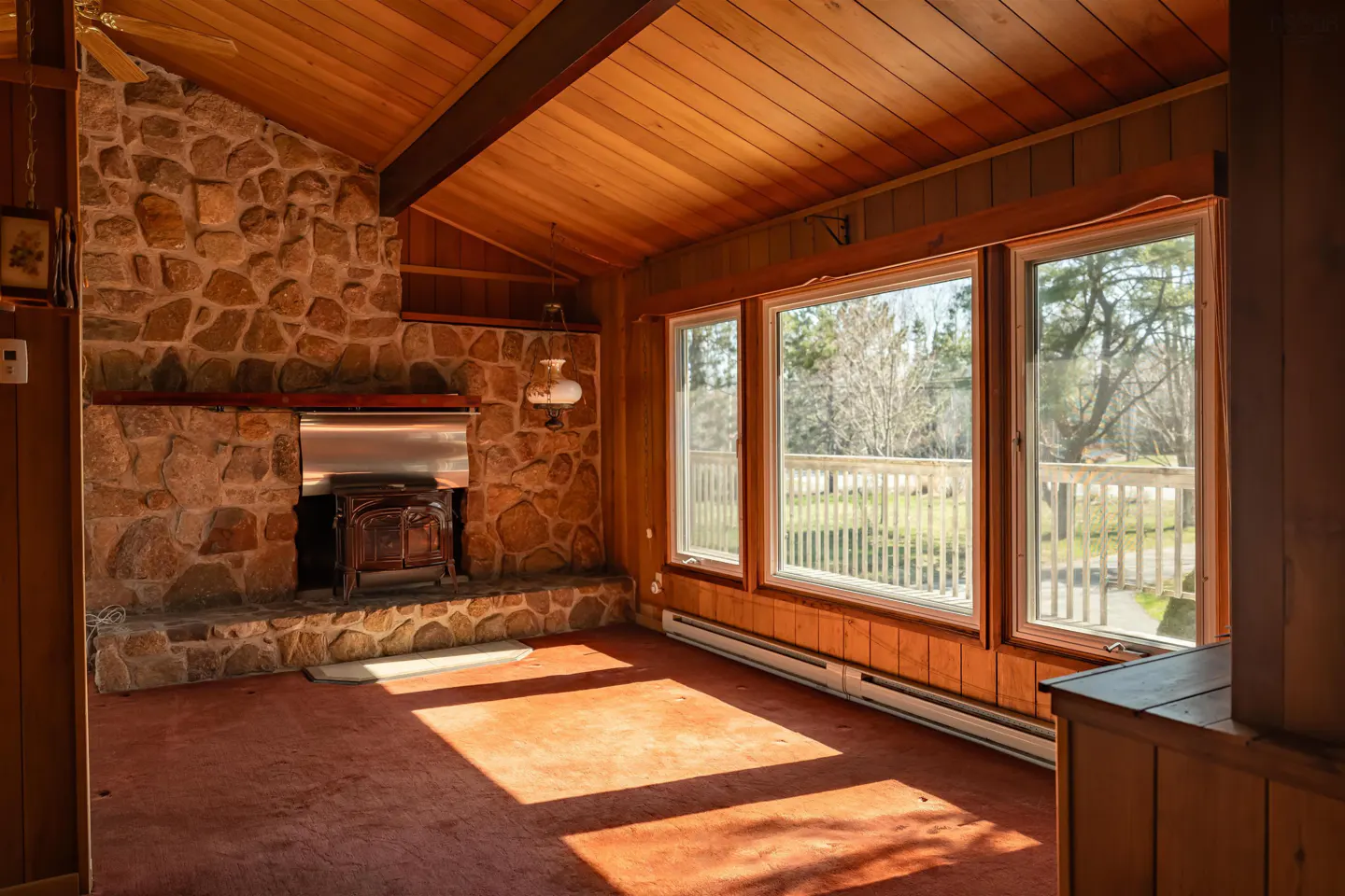 A living room with wood paneling, a stone fireplace with a wood-burning stove, and three large windows overlooking a deck and trees.
