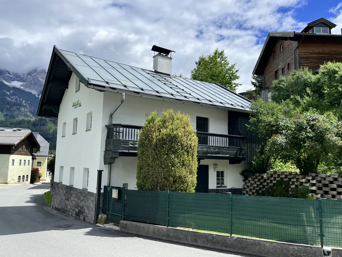 White two-story house with a metal roof, dark balcony, and green fence, set against a backdrop of mountains and trees.