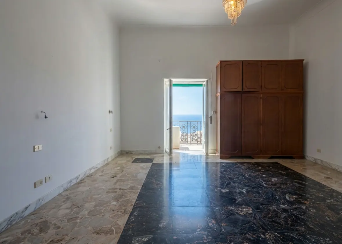 Empty room with marble floors, white walls, and a wooden wardrobe. Balcony doors open to a sea view. Chandelier hangs from the ceiling.