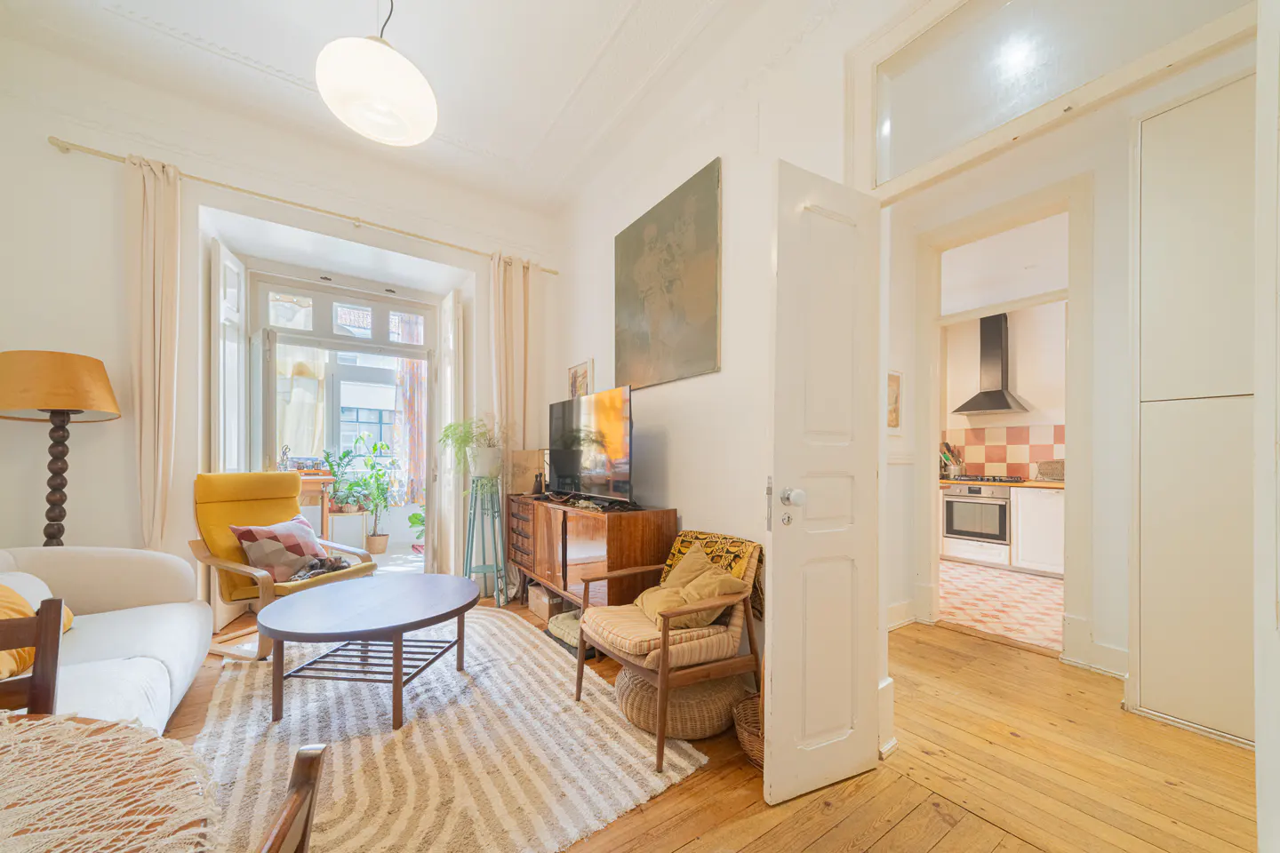 Bright living room with a yellow chair, white sofa, and oval coffee table on a striped rug. A doorway leads to a kitchen with checkered tiles.