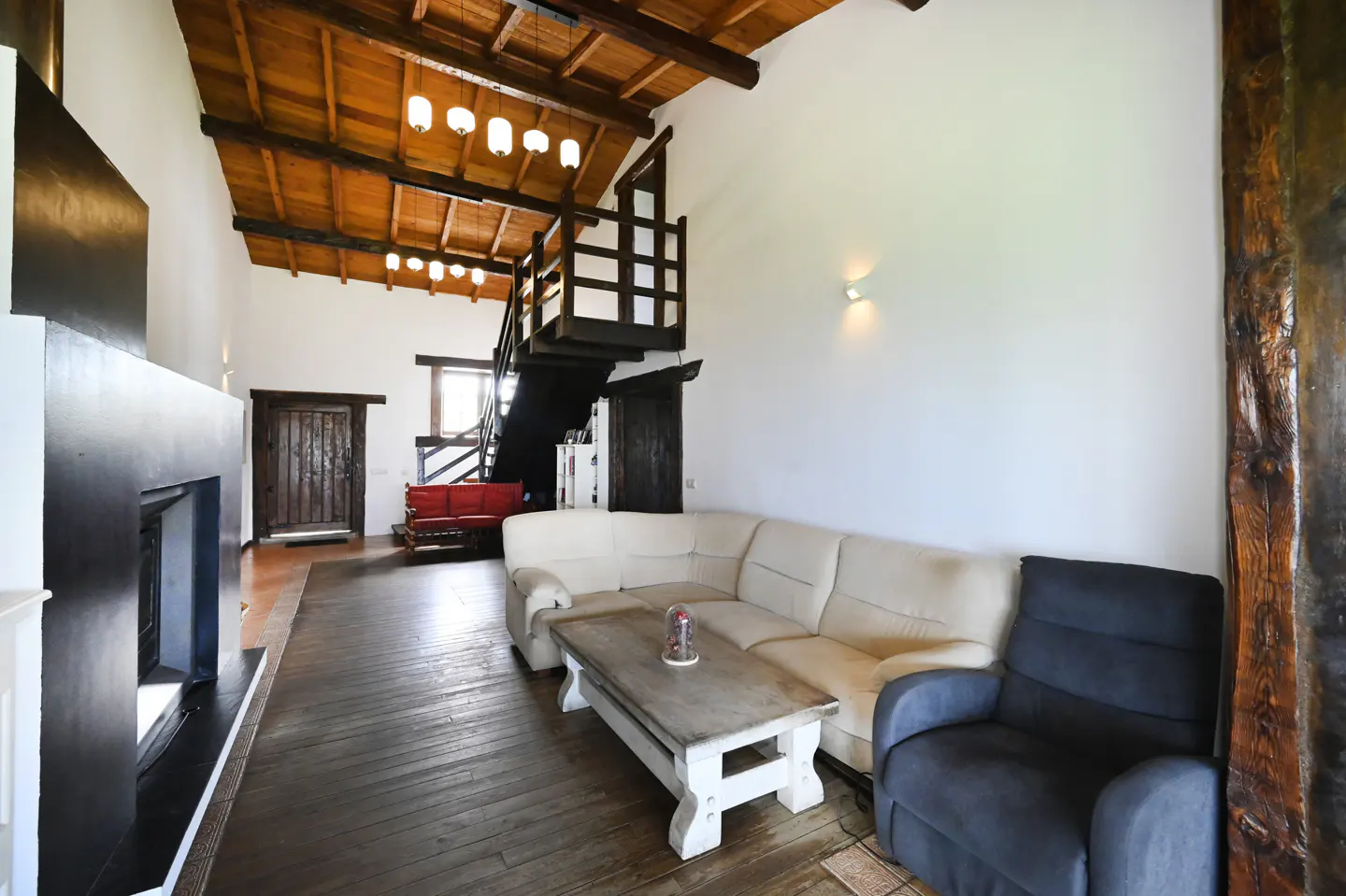 Living room with white walls, wood floors, and a high, wood-beamed ceiling. A white sofa, blue armchair, and wood coffee table sit on the floor. A black fireplace is on the left.