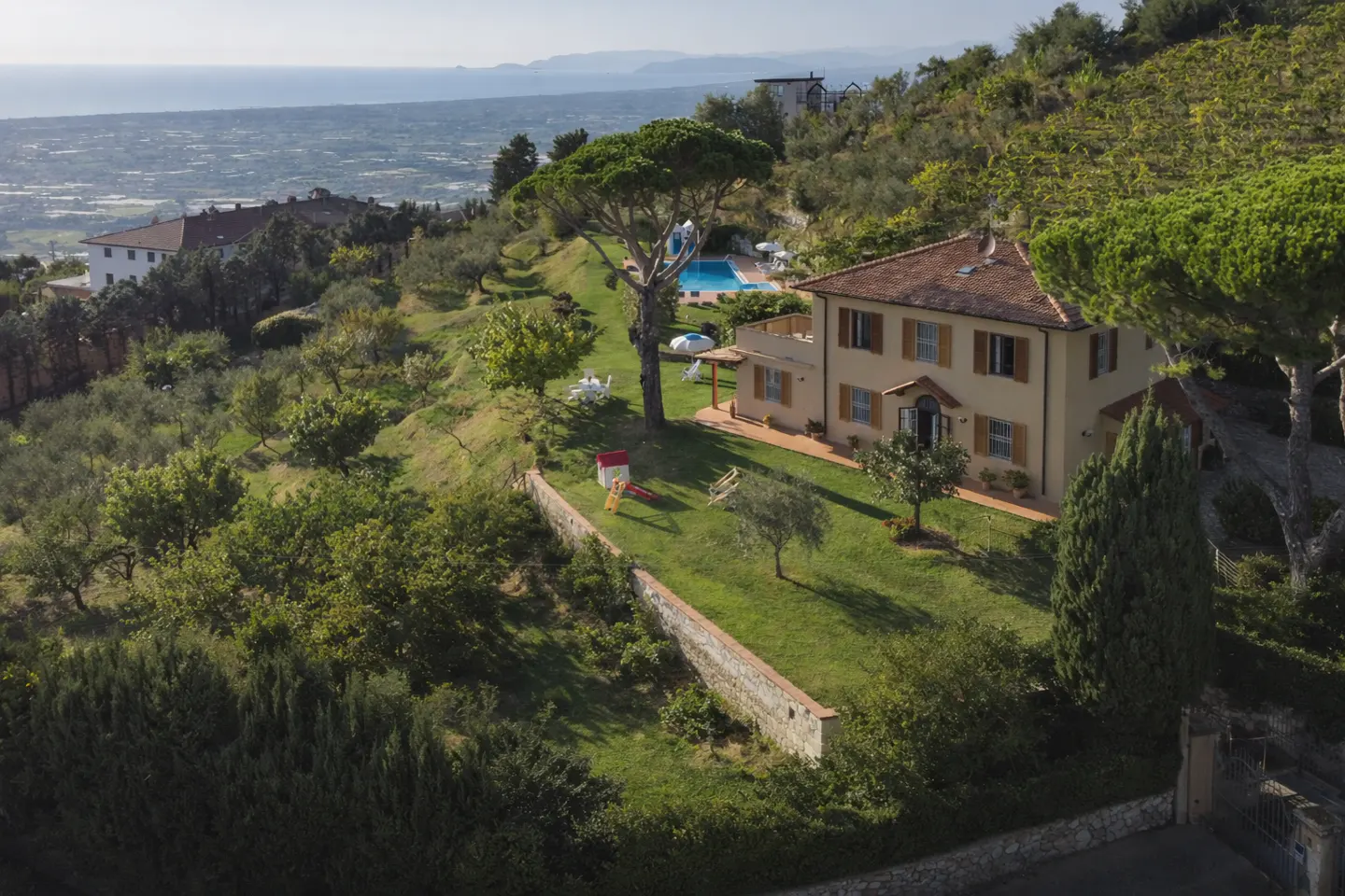 Aerial view of a two-story beige house with a red tile roof on a green hillside with a pool and ocean view.