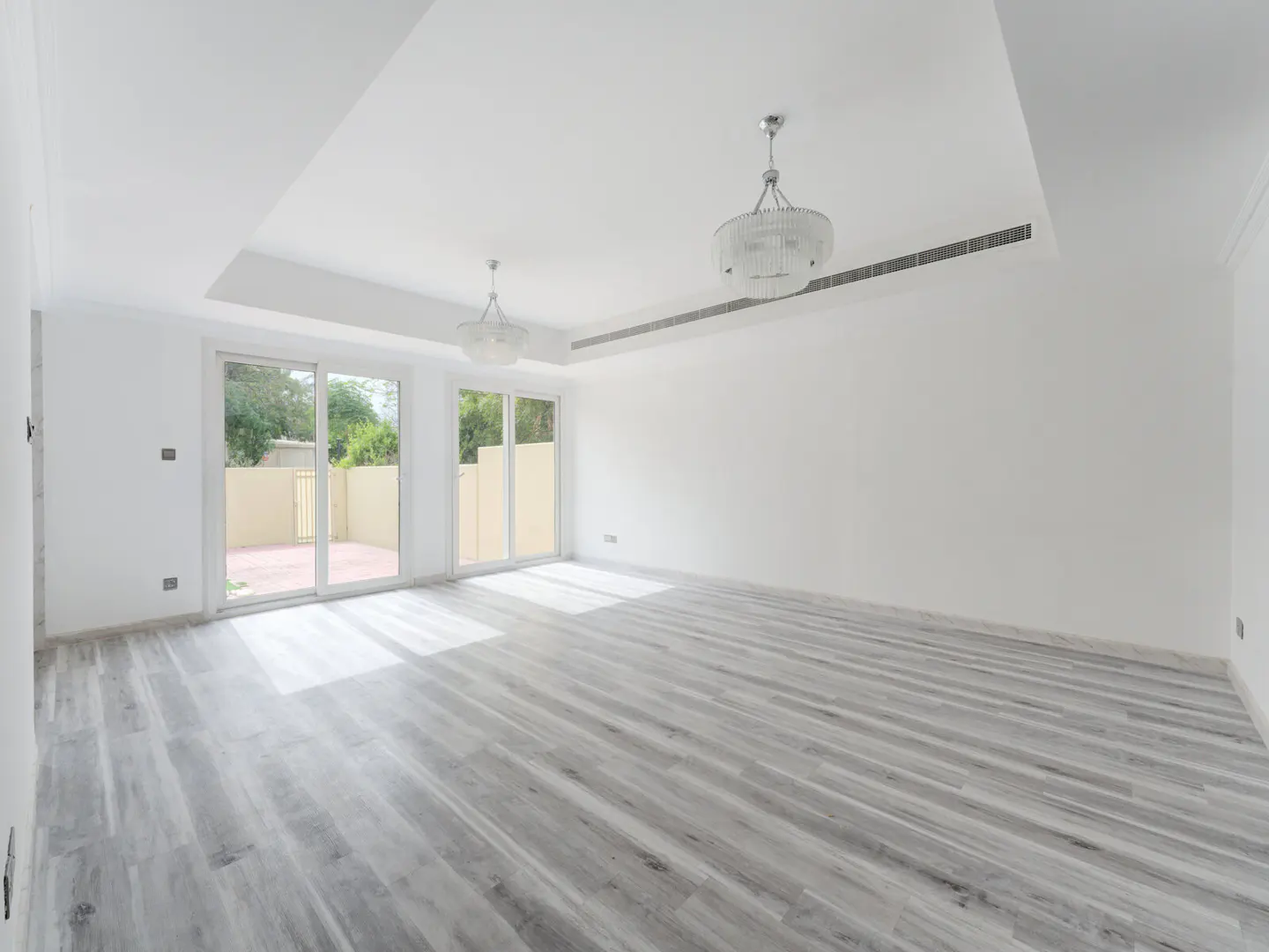 Bright, empty room with gray wood floors, white walls, and two chandeliers. Sliding glass doors lead to a patio.