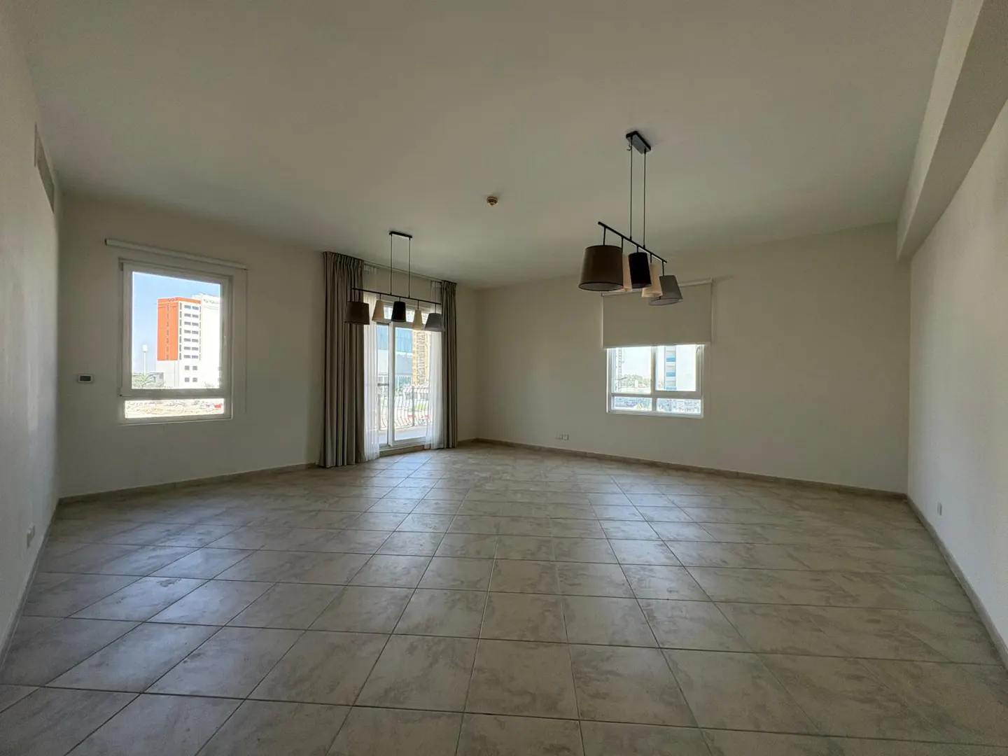 An empty, bright room with beige tile flooring, white walls, two windows, and a sliding glass door with curtains. Two modern chandeliers hang from the ceiling.