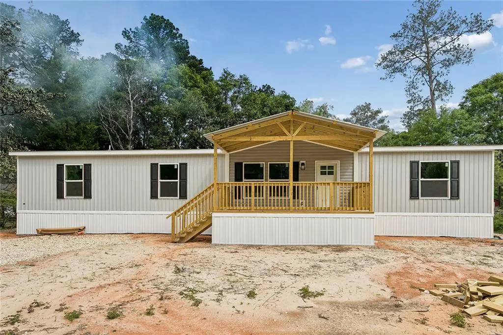 Exterior view of a gray mobile home with a wooden porch and black shutters, surrounded by trees.