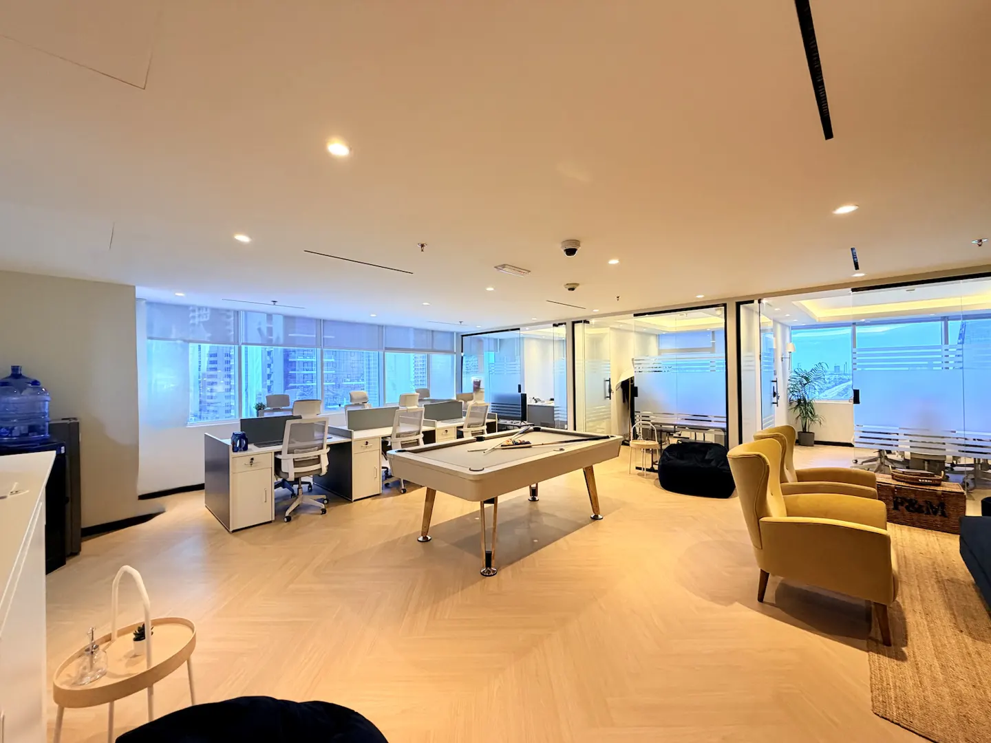 Modern office lounge with a pool table, yellow armchairs, workstations, and light wood floors with city views.
