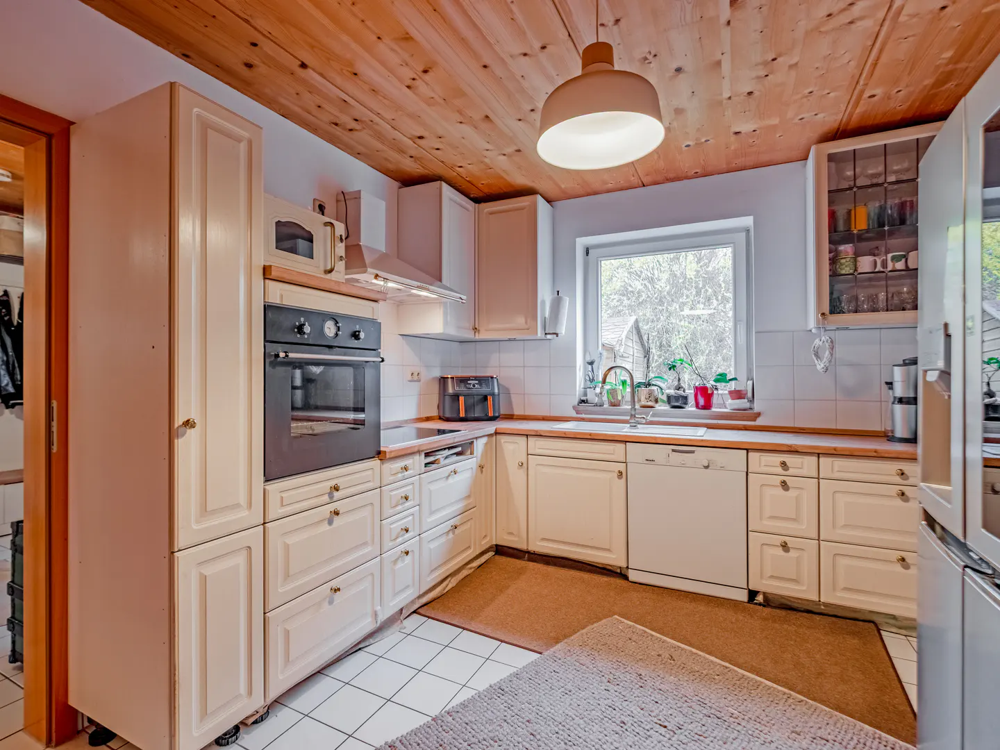 Bright kitchen with white cabinets, black oven, and wood ceiling. A window overlooks greenery. A rug is on the tiled floor.