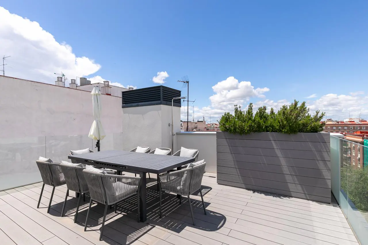 Outdoor patio with gray table, eight chairs, and a closed white umbrella. A planter box with green plants sits near a glass railing. Blue sky with clouds.