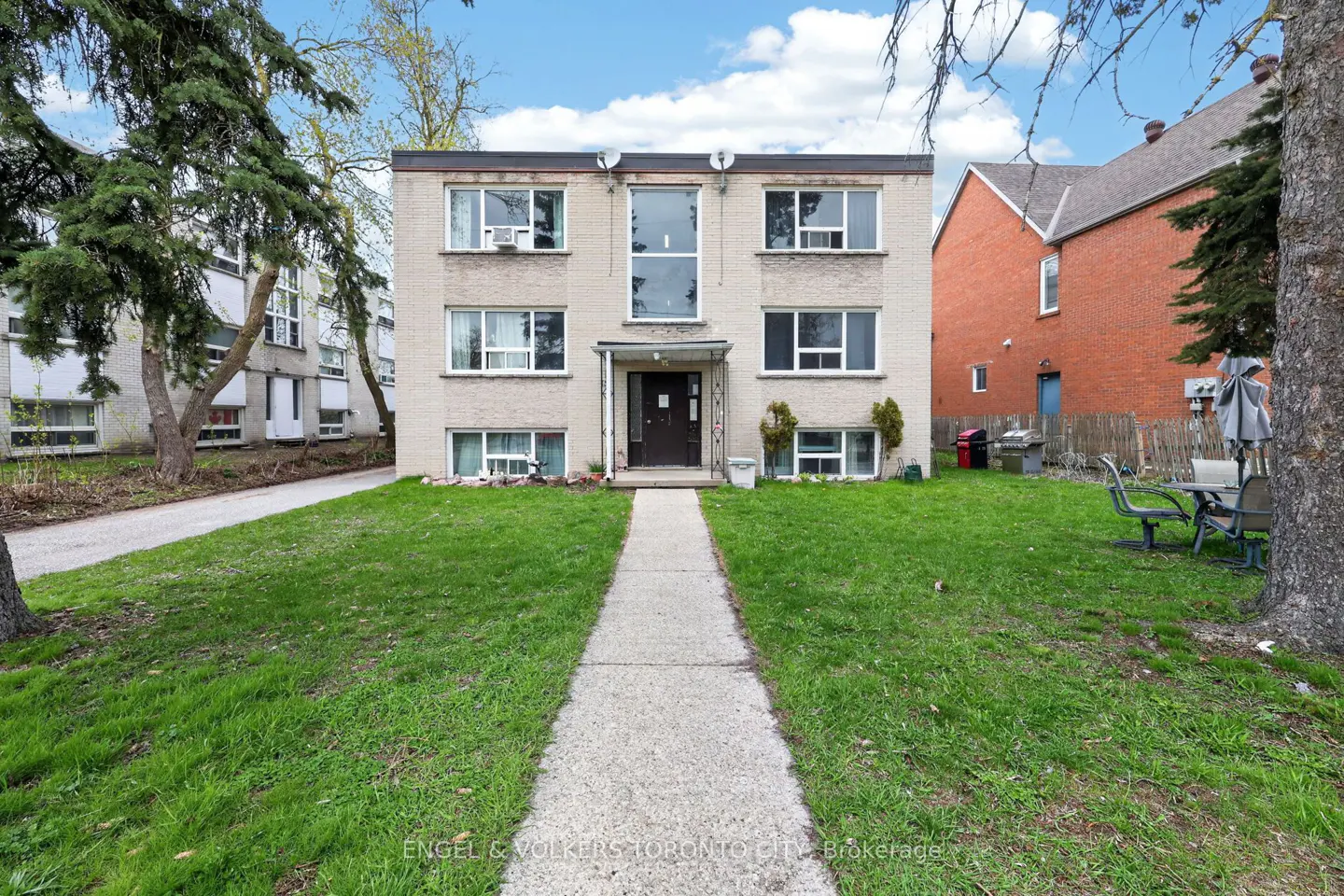 Two-story apartment building with a gray brick facade, a dark door, and a concrete walkway leading to it. Green lawn and trees surround the building.