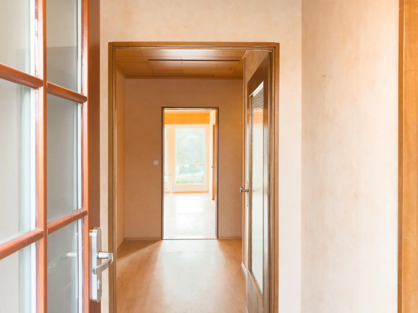 Hallway view with wood floors, peach walls, and open wood-framed doors leading to a bright room with a window.