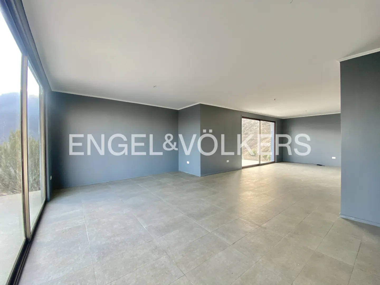 An empty room with gray walls, tile floors, and large windows with a view. Engel & Völkers logo is visible.