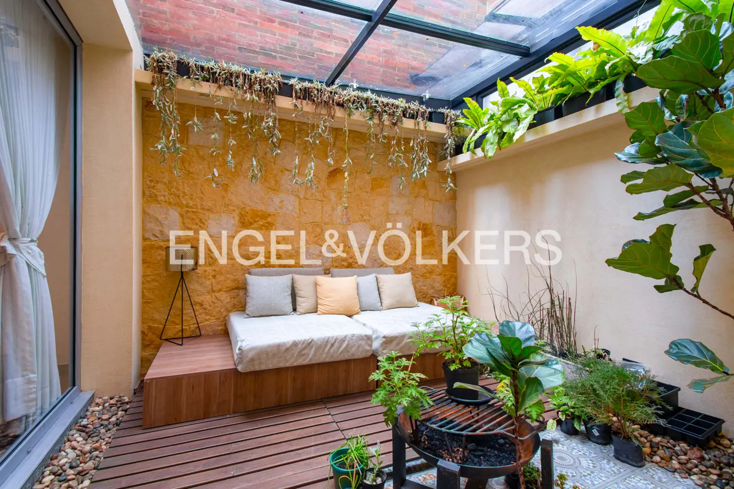 Indoor patio with stone wall, wooden deck, and glass roof. A white couch with pillows sits against the wall, surrounded by plants.