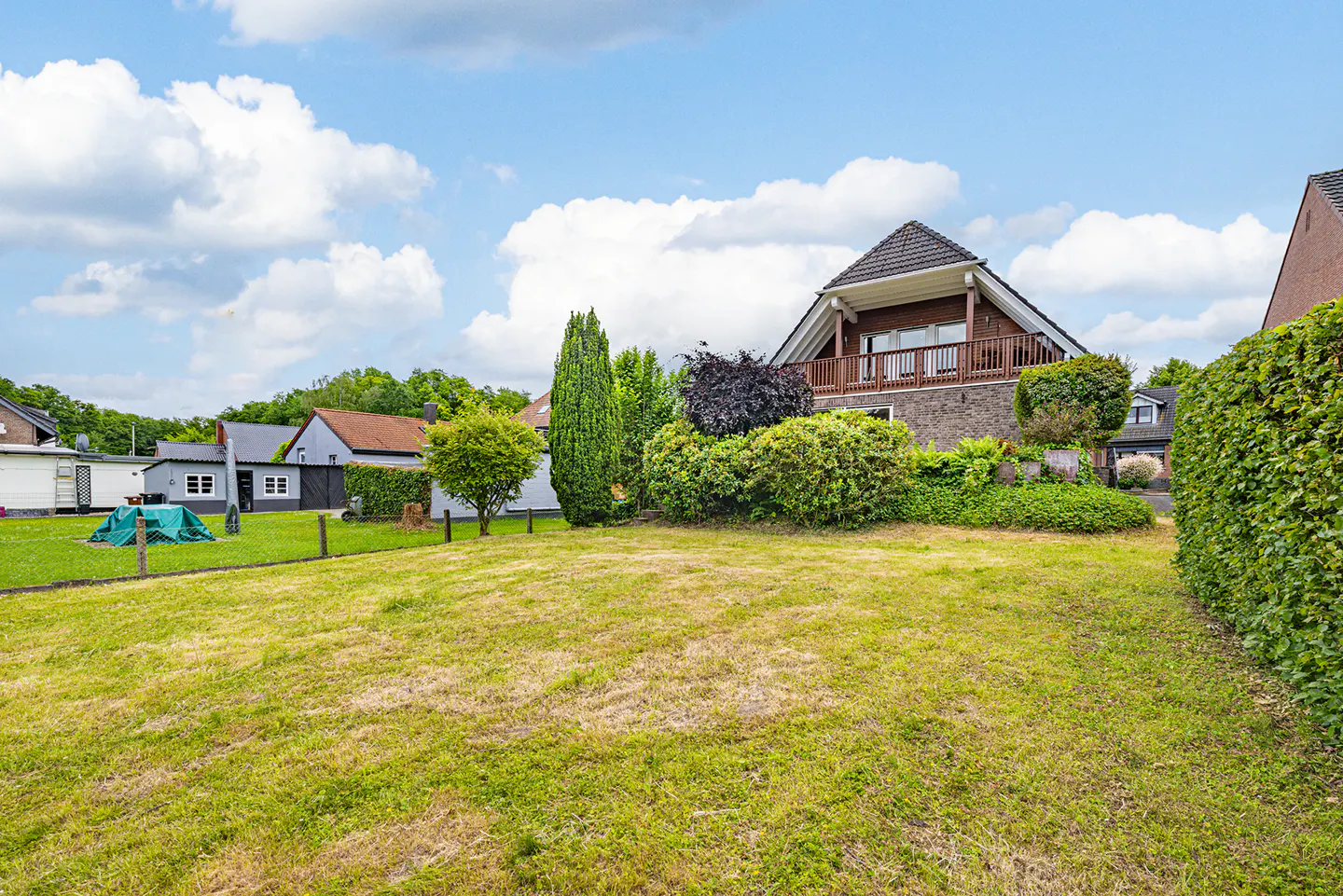 A large, grassy backyard with a stone house featuring a wooden balcony, surrounded by lush green hedges and trees.