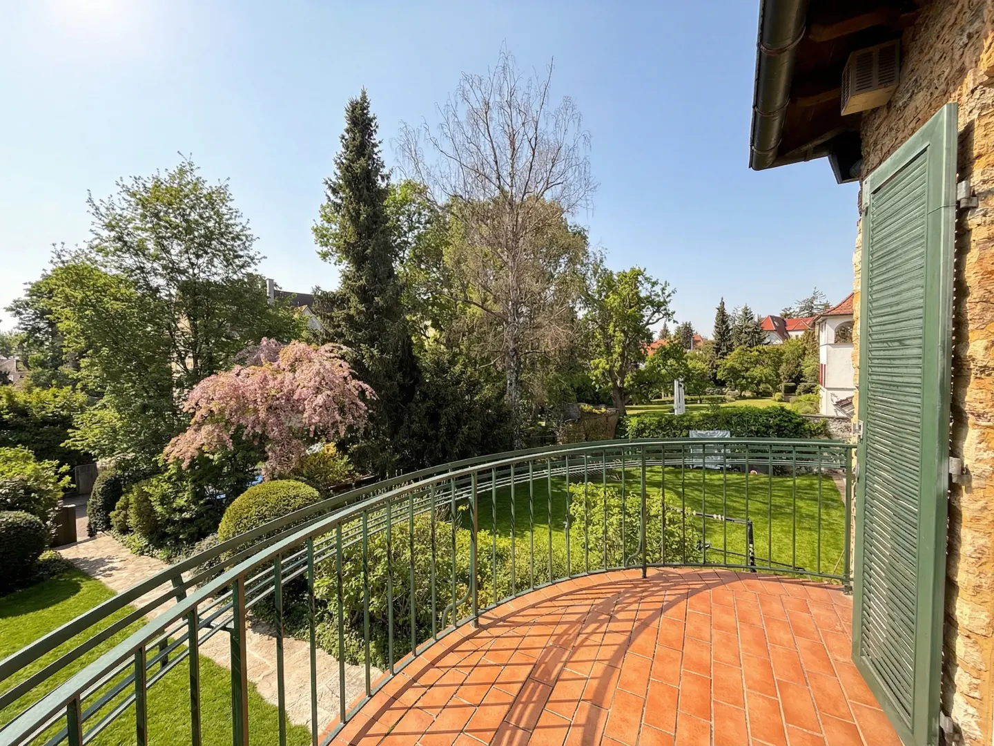 View from a balcony with red tile floor and green railing, overlooking a lush green garden with trees and a blue sky.