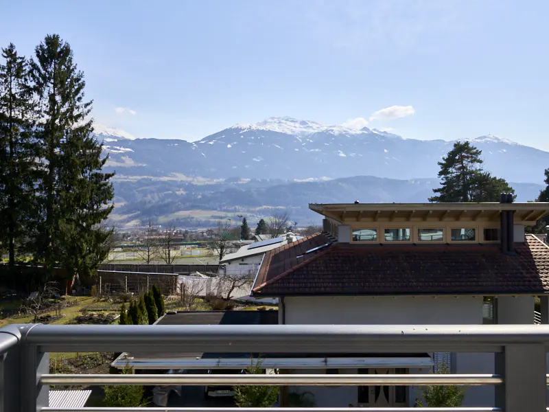 View from a balcony overlooking a red-roofed house, trees, and snow-capped mountains under a blue sky.