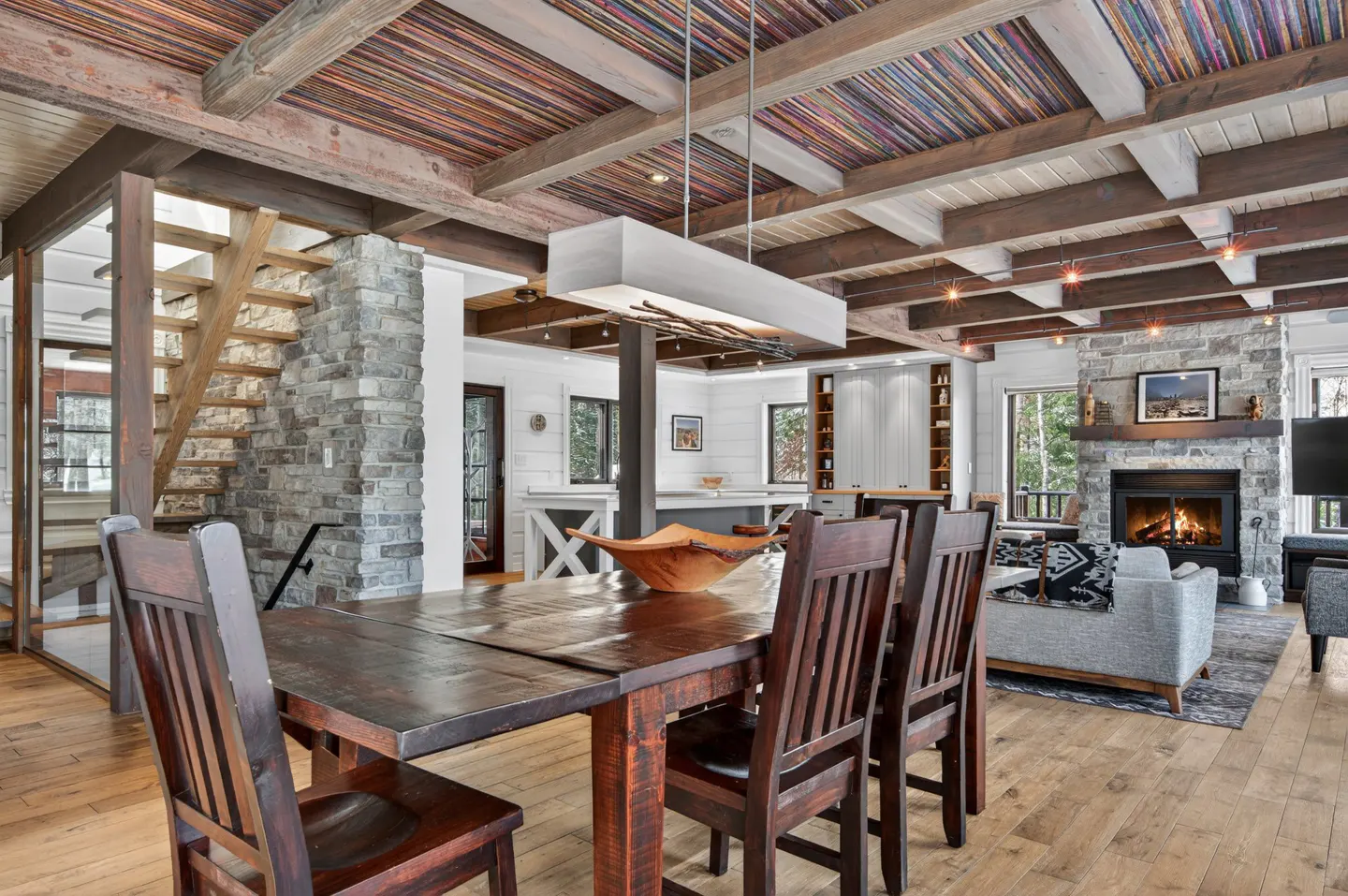 Rustic dining room with wood table, chairs, and floors. Stone fireplace and stairs. Beamed ceiling with colorful planks.