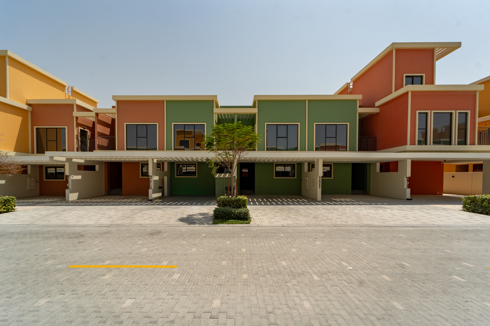 Modern townhouses with orange and green facades, featuring covered parking and a paved driveway under a clear blue sky. [AI-generated text]