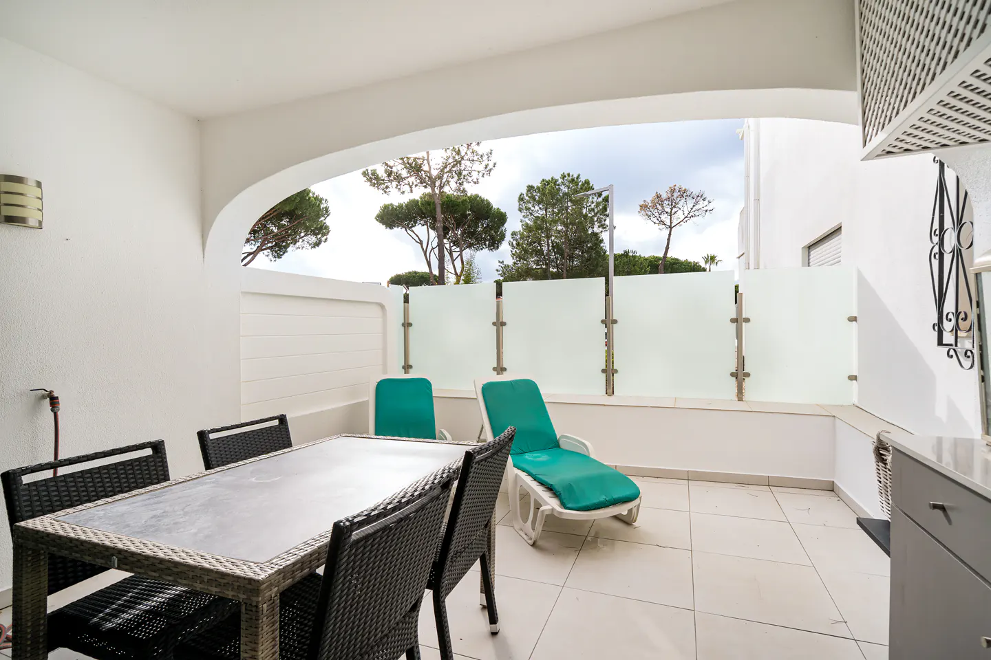 Outdoor patio with a table, chairs, and lounge chairs. White walls and tile floor. Trees are visible in the background.