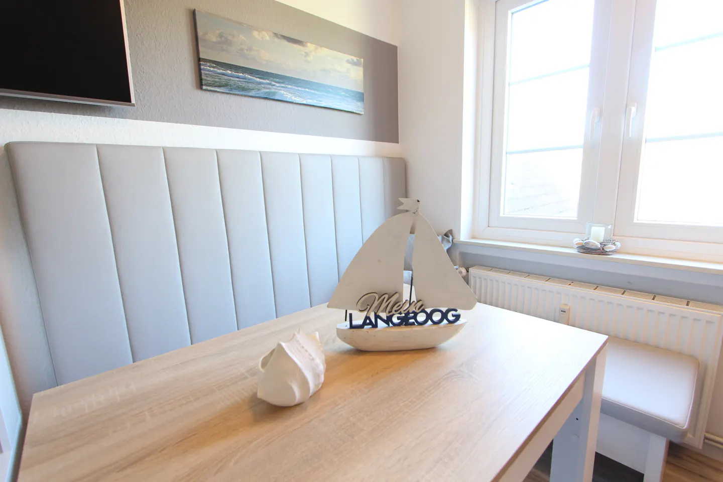 Dining area with a light wood table, white bench seating, and a decorative sailboat with "Moin Langeoog" text. A seashell sits on the table. A window provides natural light.