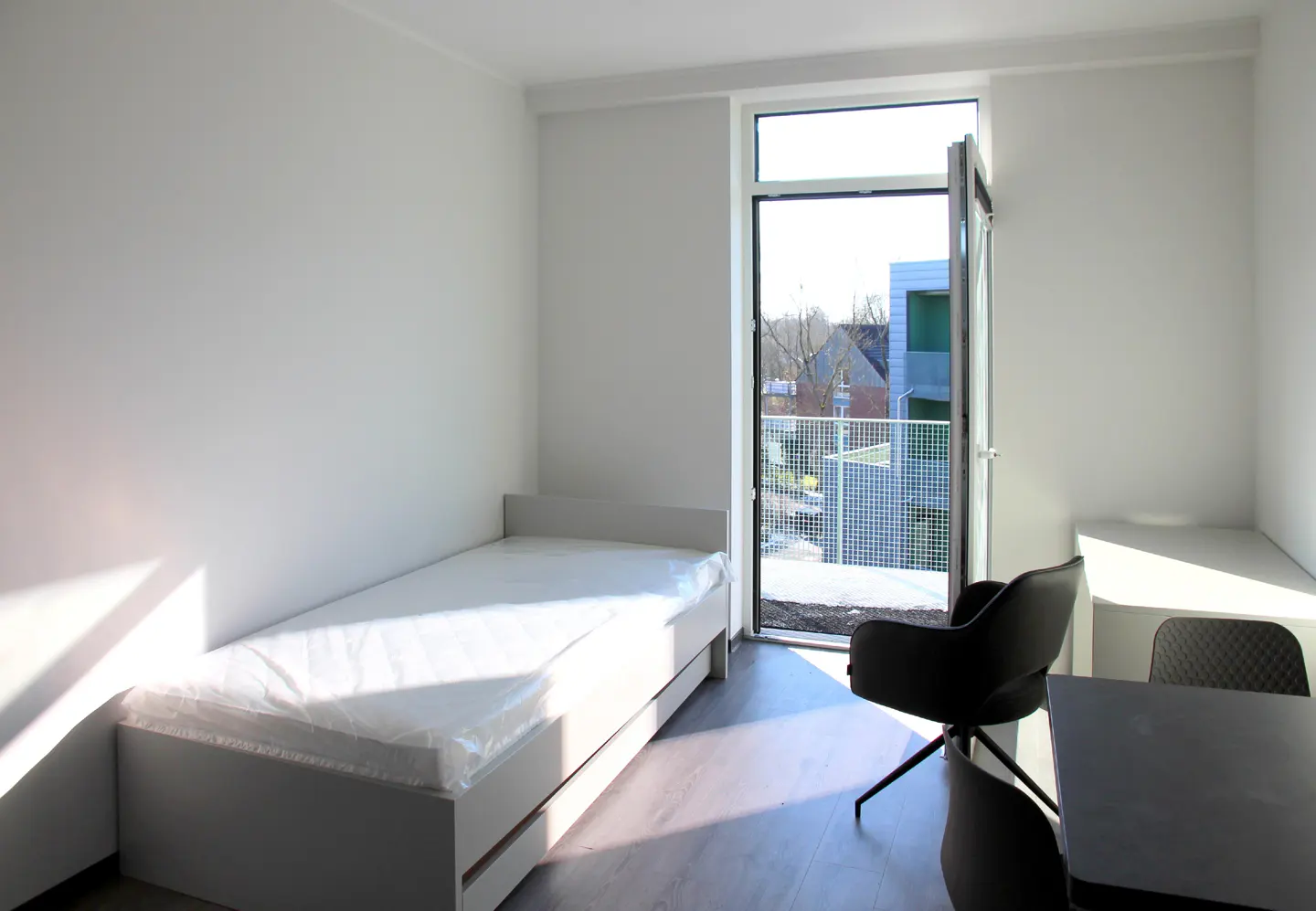 Bright, modern room with a single bed still wrapped in plastic, a desk, and a black chair. A door opens to a balcony with a view of nearby buildings.