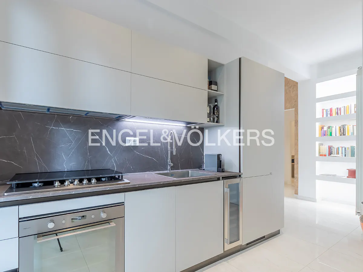 Modern kitchen with light gray cabinets, dark marble backsplash, stainless steel oven, and a built-in bookshelf nearby.