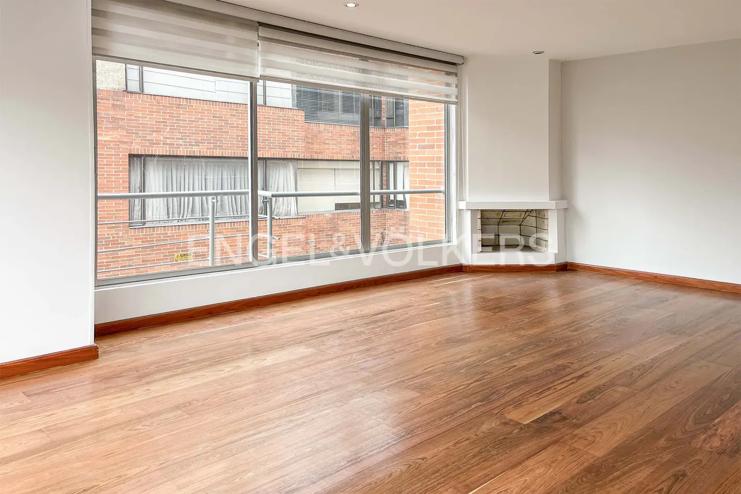 Empty room with hardwood floors, white walls, and a large window with blinds. A white fireplace is on the right wall.