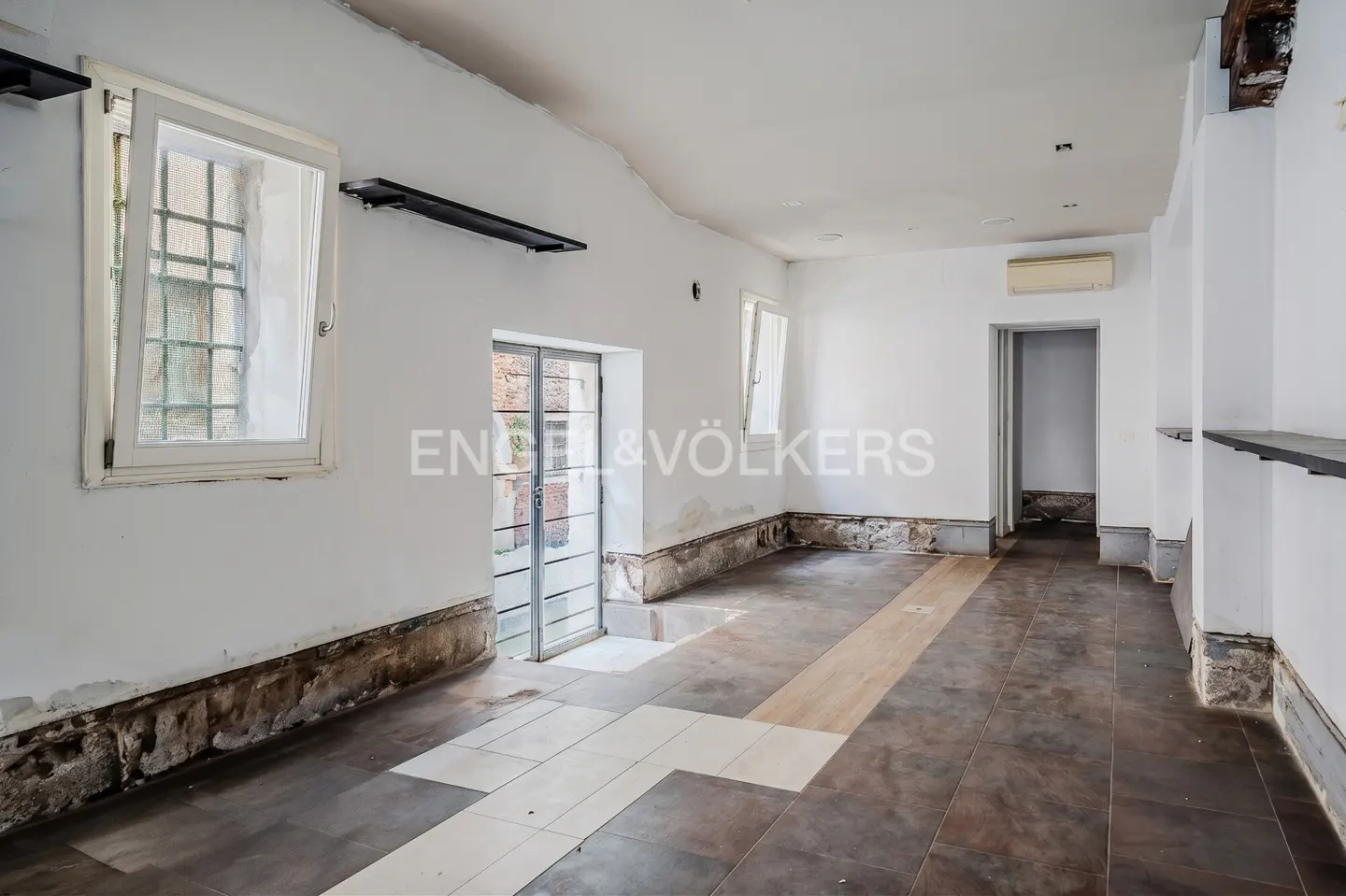 Unfurnished room with white walls, brown tile floor, and windows. Walls show signs of wear. Doorway leads to another room.