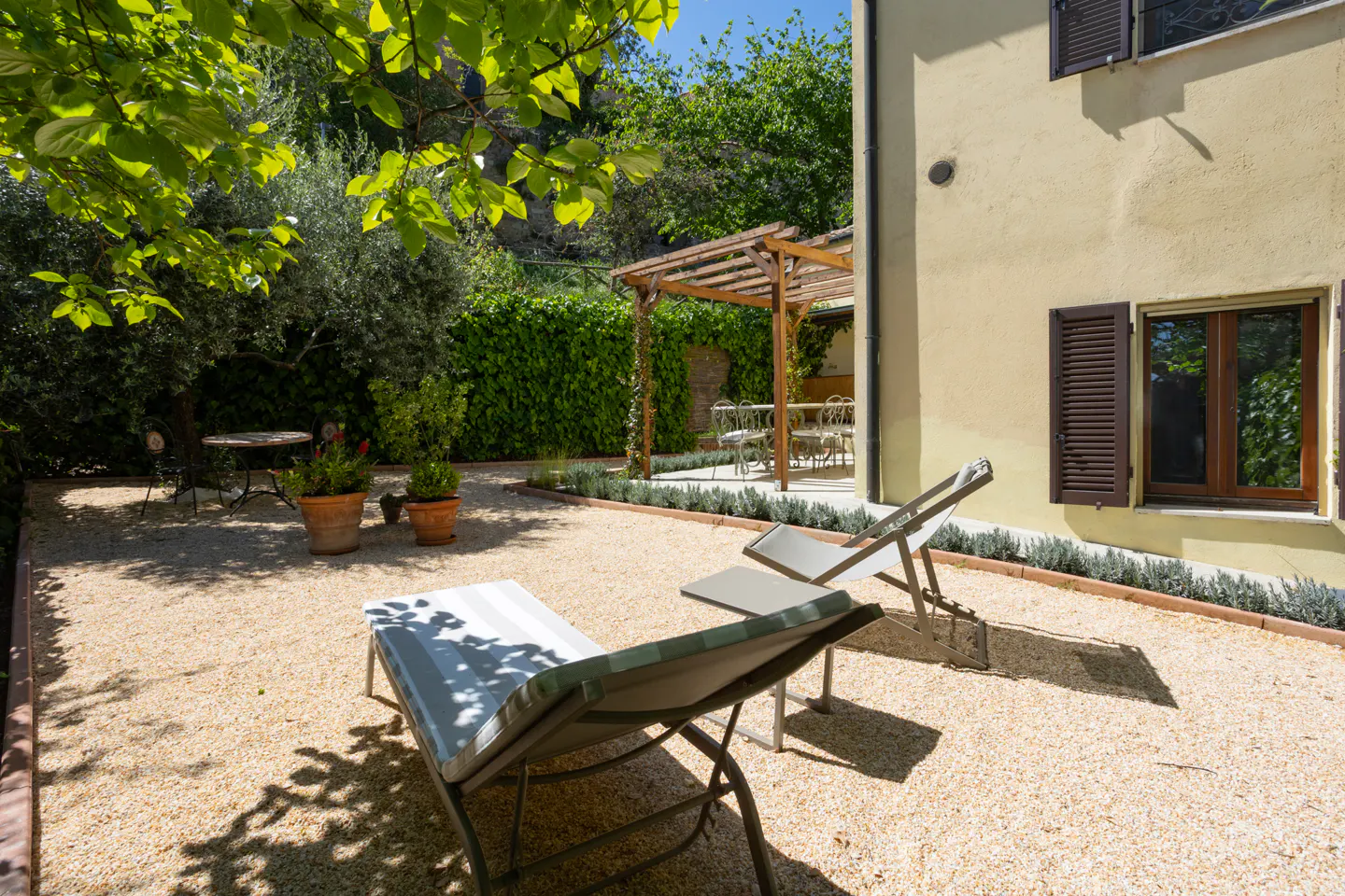 Outdoor patio with two lounge chairs on a gravel surface, a wooden pergola, and a yellow building with brown shutters.