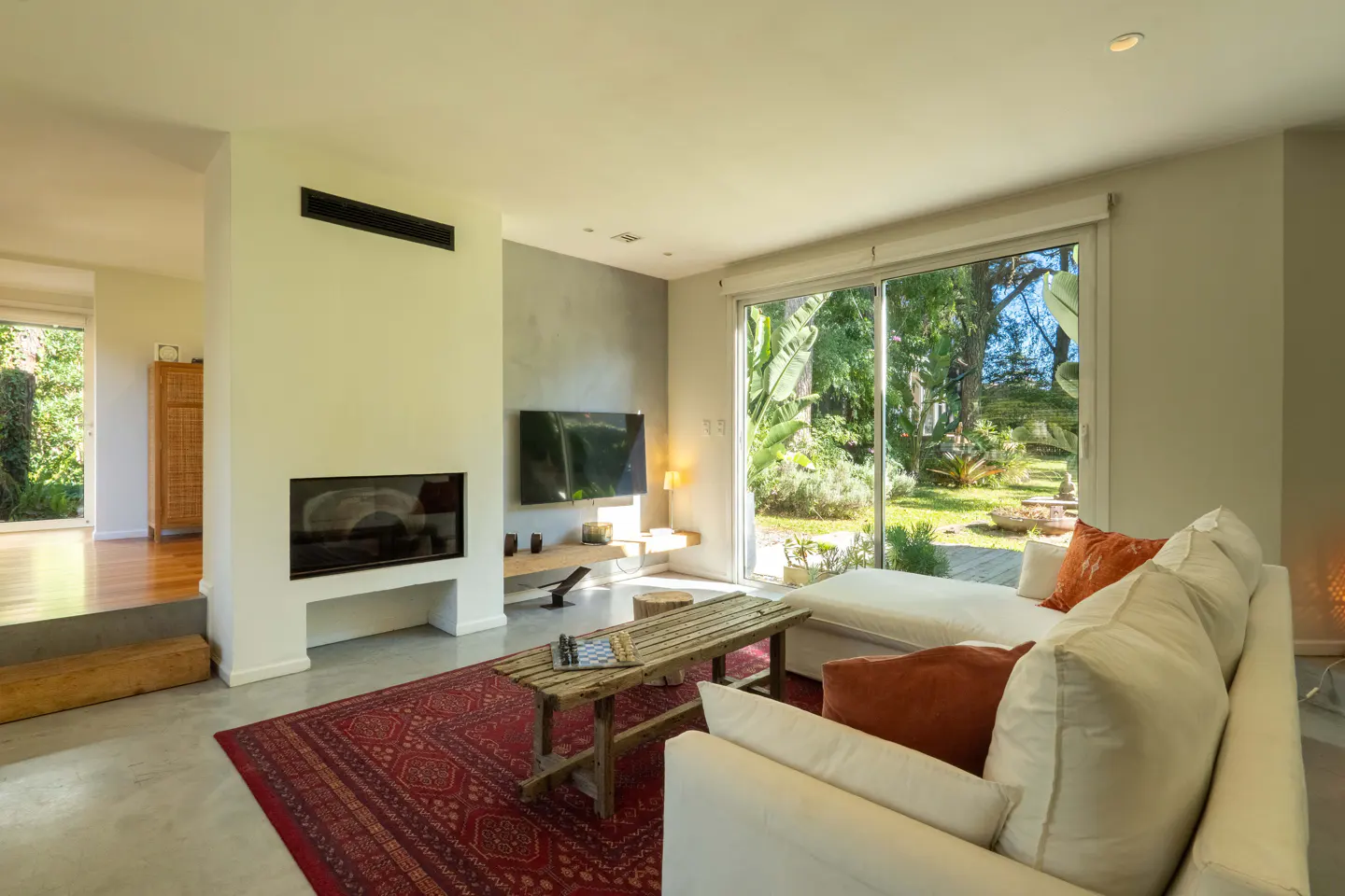 Bright living room with white sofa, rustic wood table on red rug, fireplace, and large window view of lush green garden.