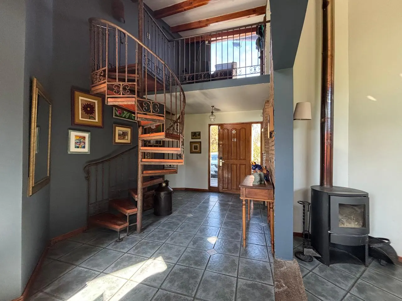 Interior view of a home's entrance with a spiral staircase, wood-burning stove, and tiled floor. The walls are gray and cream.