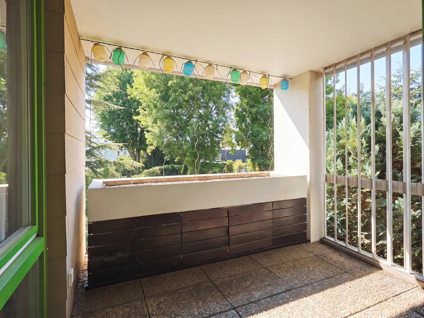 A balcony with a view of trees. The balcony has a brown wood panel, a concrete floor, and colorful lights strung above.