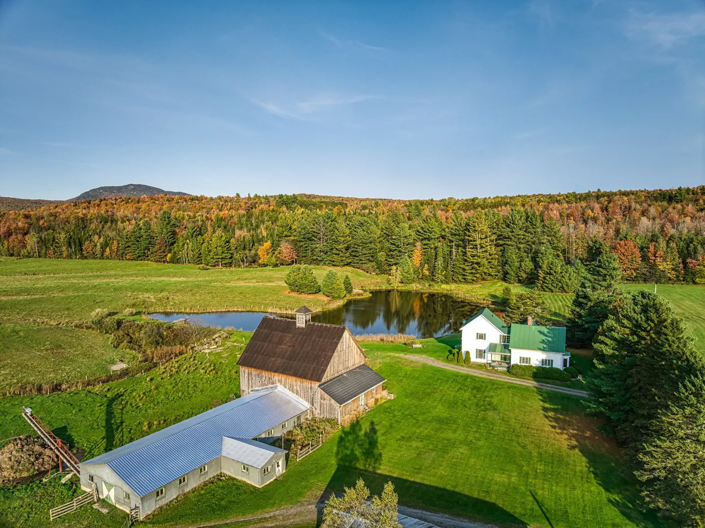 Aerial view of a farm with a barn, pond, and white house with a green roof, surrounded by green fields and colorful autumn trees.