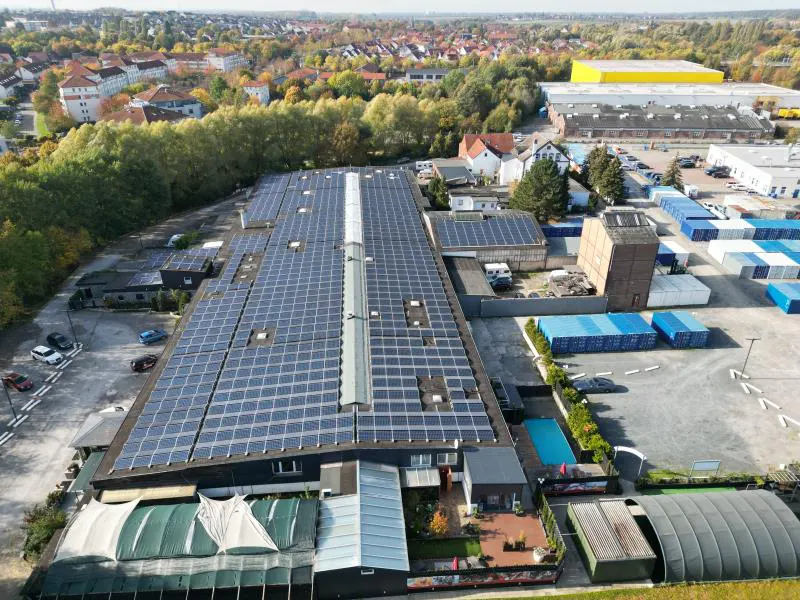 Aerial view of a large, flat-roofed commercial building covered in solar panels, surrounded by trees and other buildings.