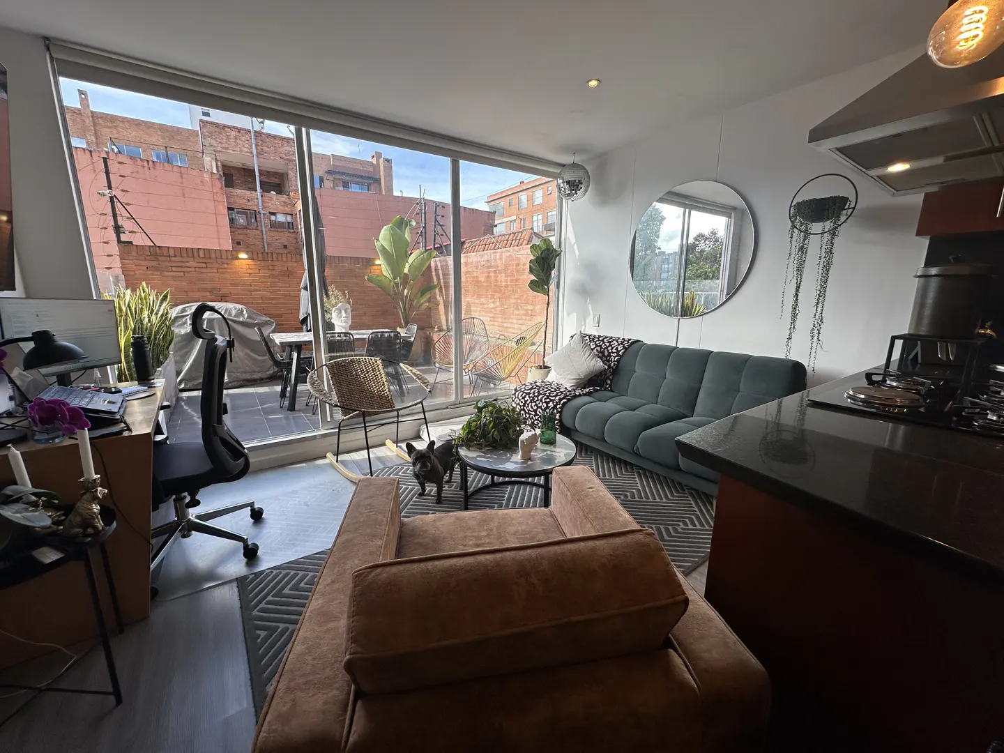 Modern apartment living room with a brown armchair, green sofa, desk, and a patio view through large glass sliding doors.