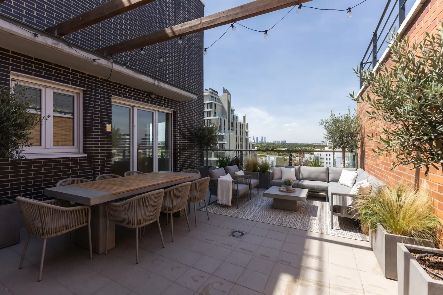 Rooftop patio with dining table, gray wicker seating, and city views. String lights hang from a wooden pergola. Brick walls and potted plants add warmth.
