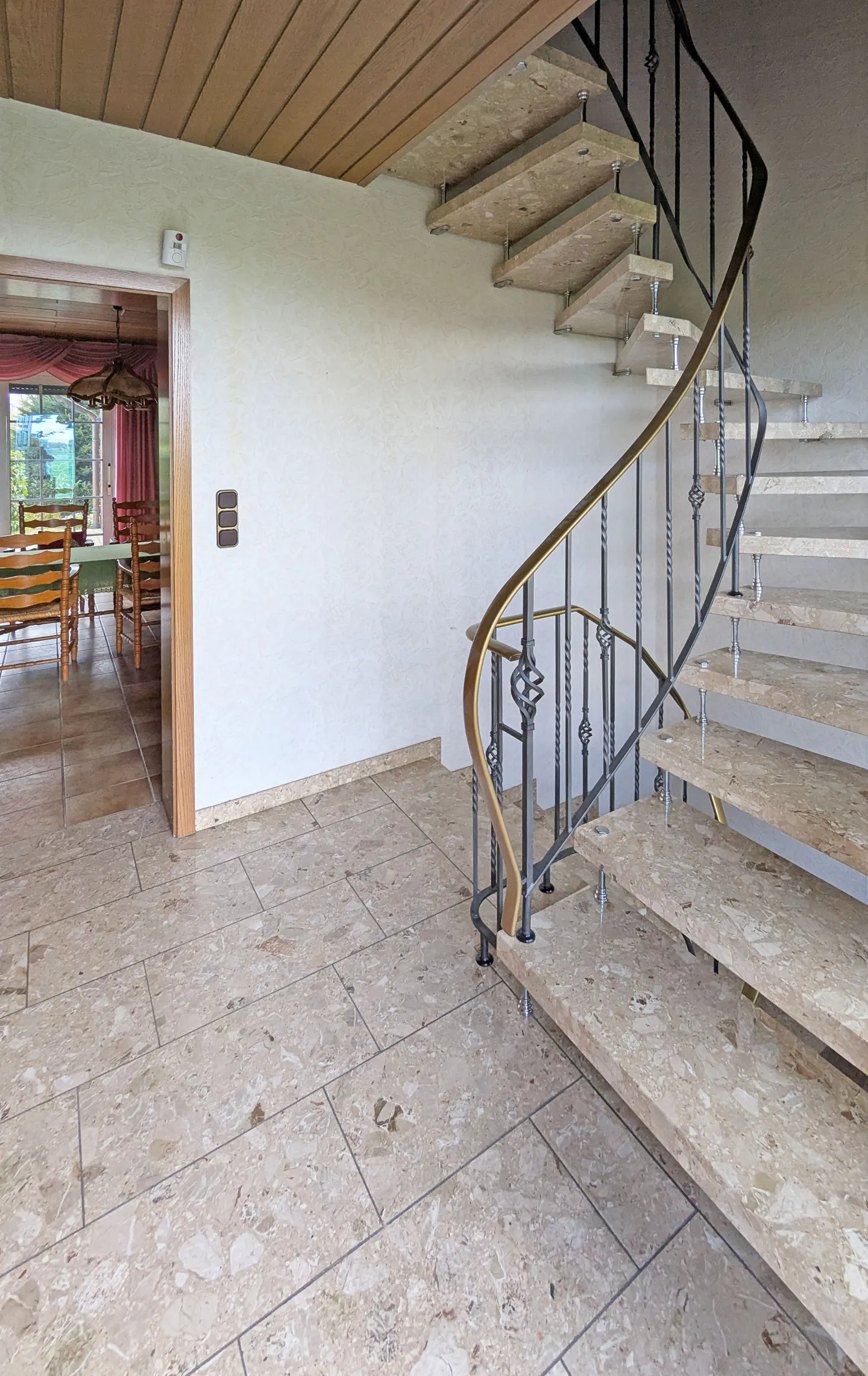 Interior view of a foyer with a spiral staircase, stone floors, and a dining room visible through an open doorway.